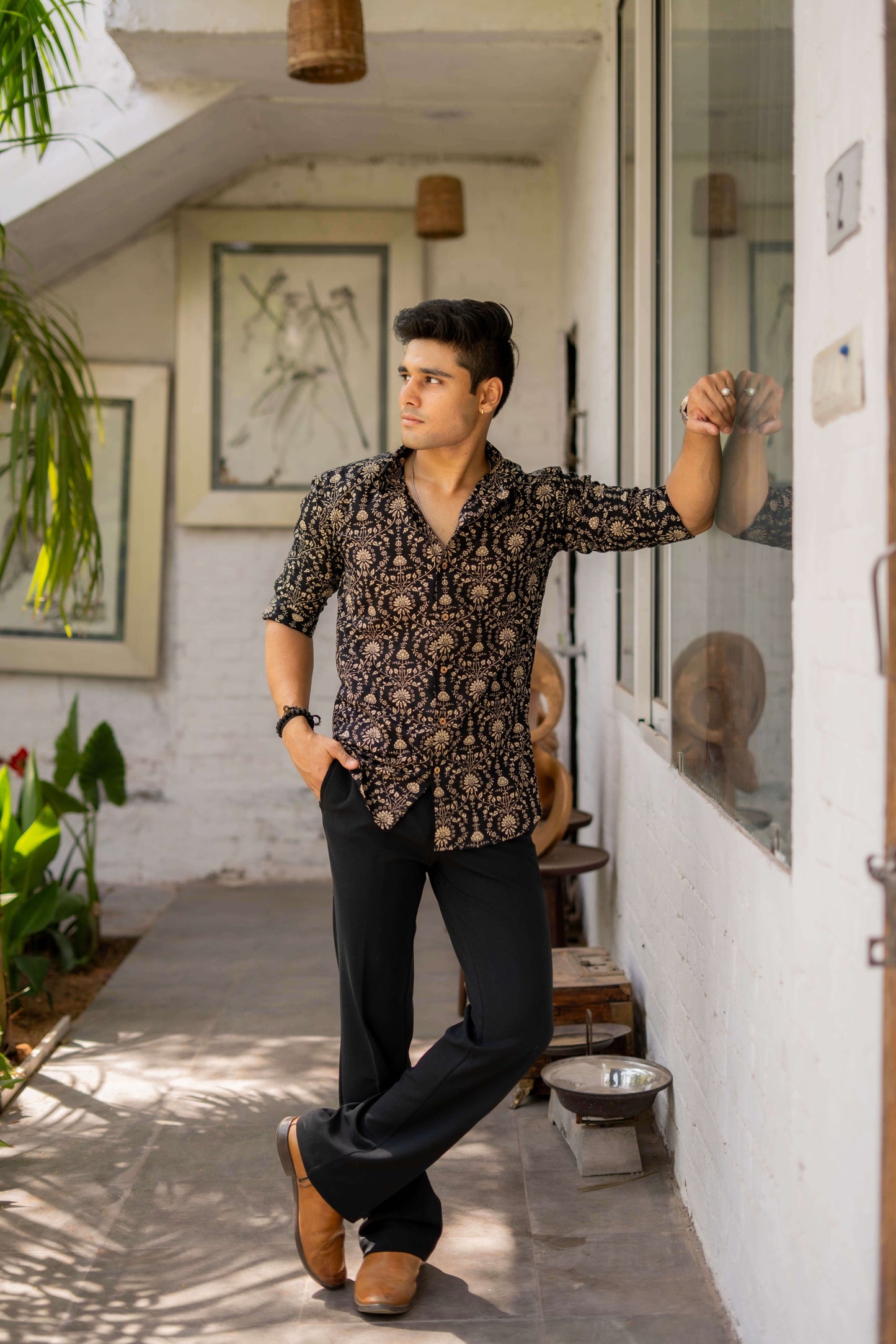 An Indian man wearing a black shirt with beige Dabu print, styling the shirt outdoors. The outfit includes a watch and a black beaded bracelet, with greenery in the background.