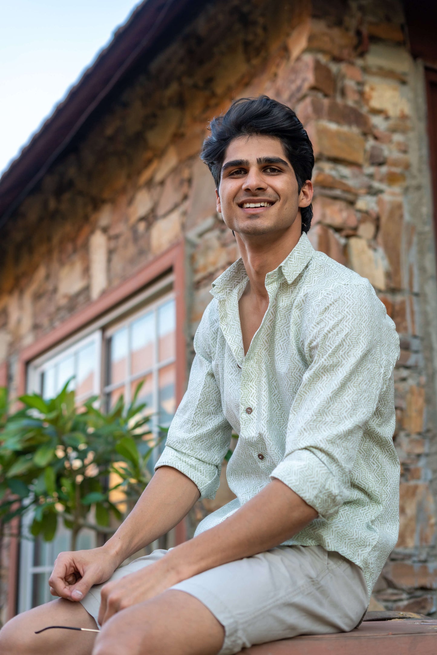 Indian man wearing a white tribal print shirt, sitting in front of a wall and a plant.