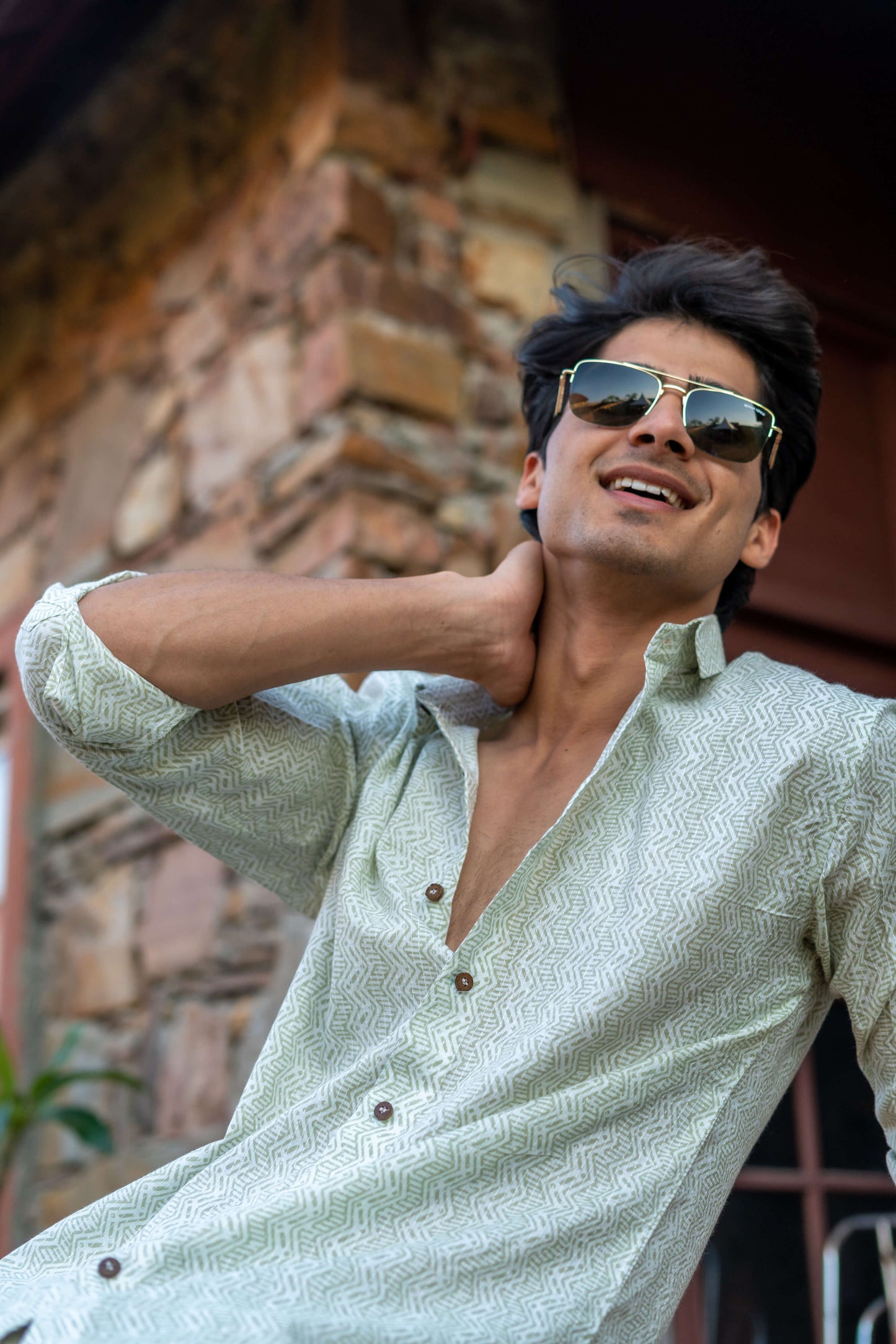 Indian Man Wearing a White Tribal Print Shirt and Black Sun Glasses, Sitting in Front of a Wall and a Plant.