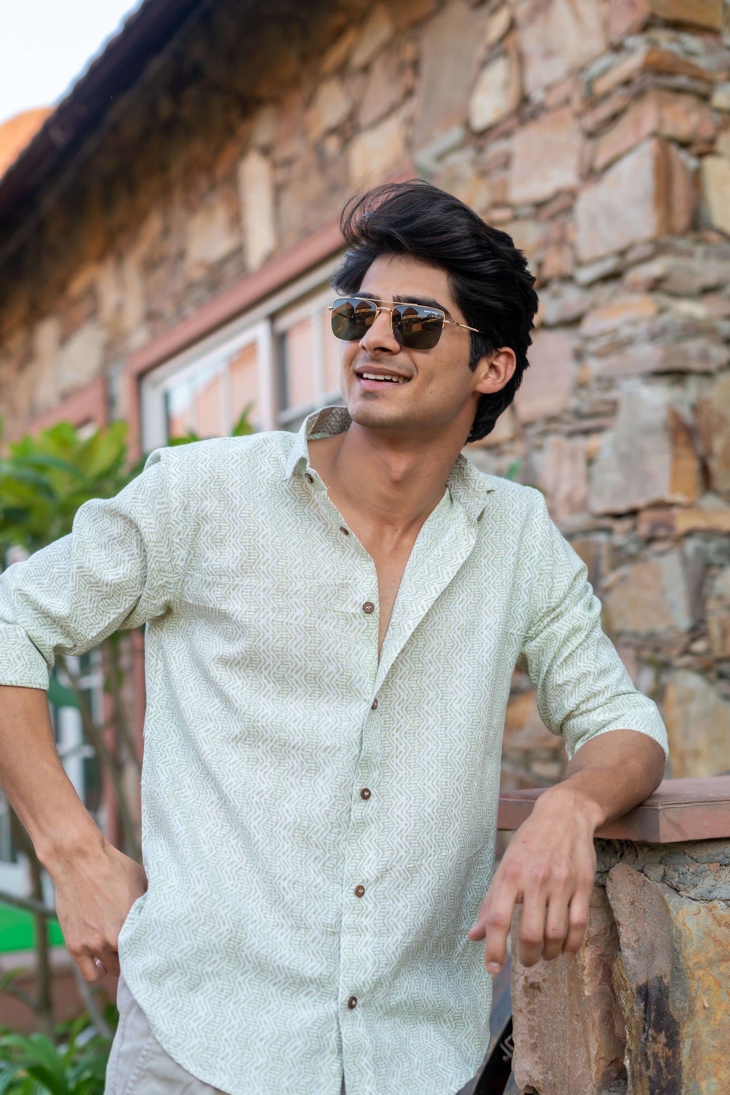Indian Man Wearing a White Tribal Print Shirt and Black Sun Glasses, Standing in Front of a Wall and a Plant.