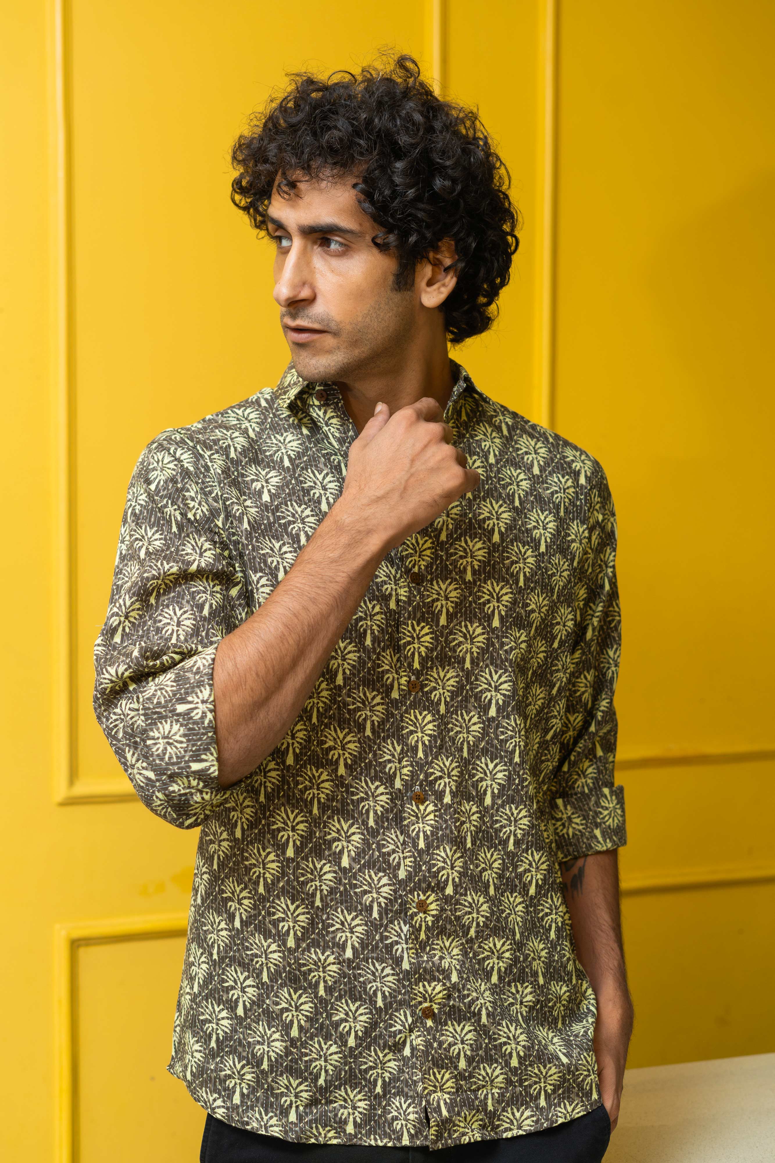 Indian man wearing a grey color shirt featuring palm tree print. standing in front of a yellow wall