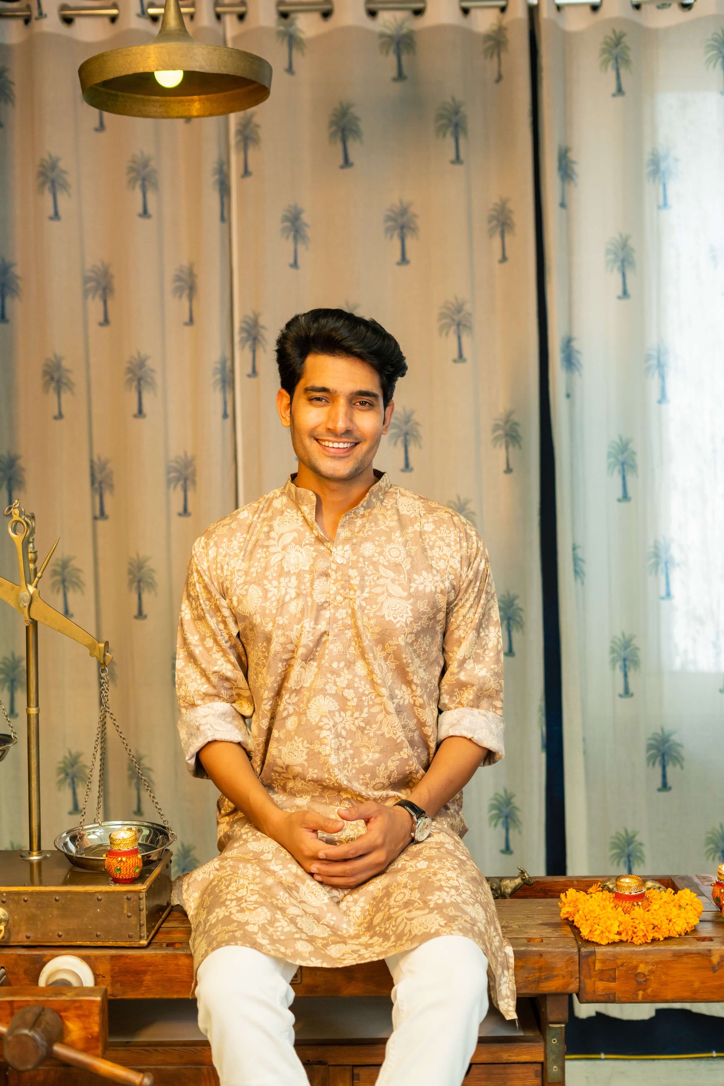 an Indian man smiling and wearing a brown color long kurta with an off-white floral print sitting on a table