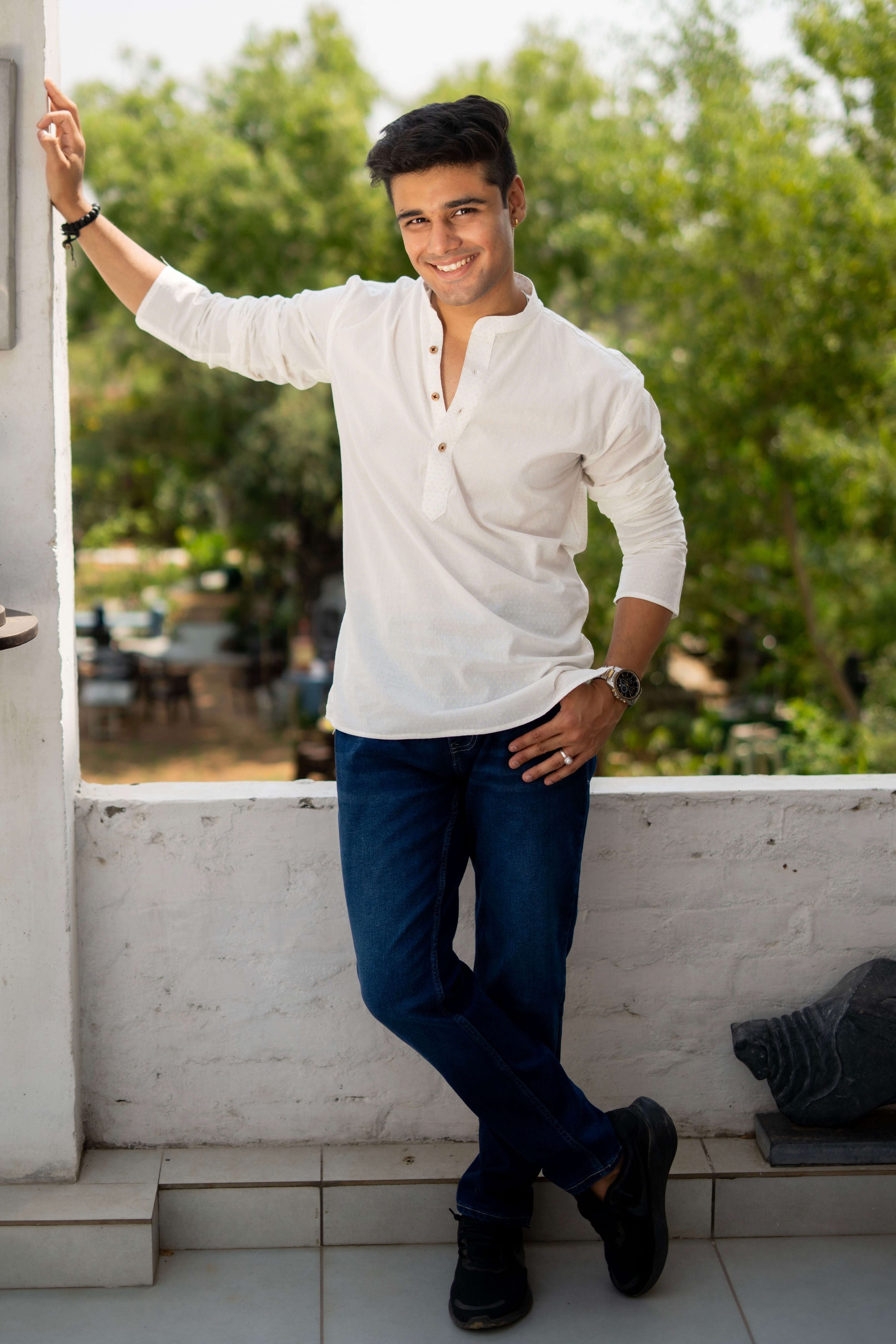 A person wearing a white short kurta with golden polka dots, paired with white pants, standing in an outdoor setting with traditional decor.