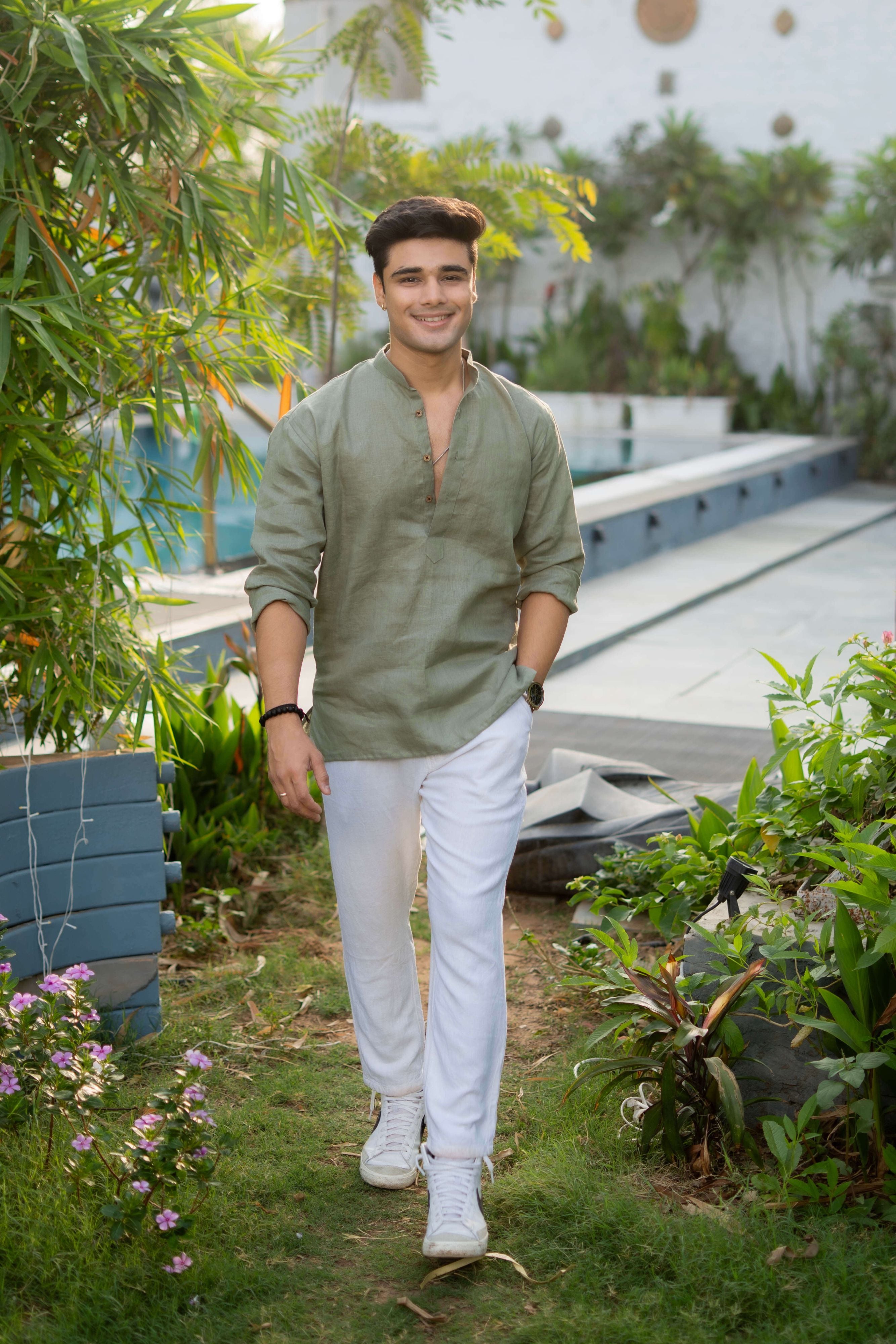 Man wearing a sage green linen short kurta with a mandarin collar and buttoned placket, paired with white cotton pants and white sneakers, walking through a garden path surrounded by greenery and flowers.