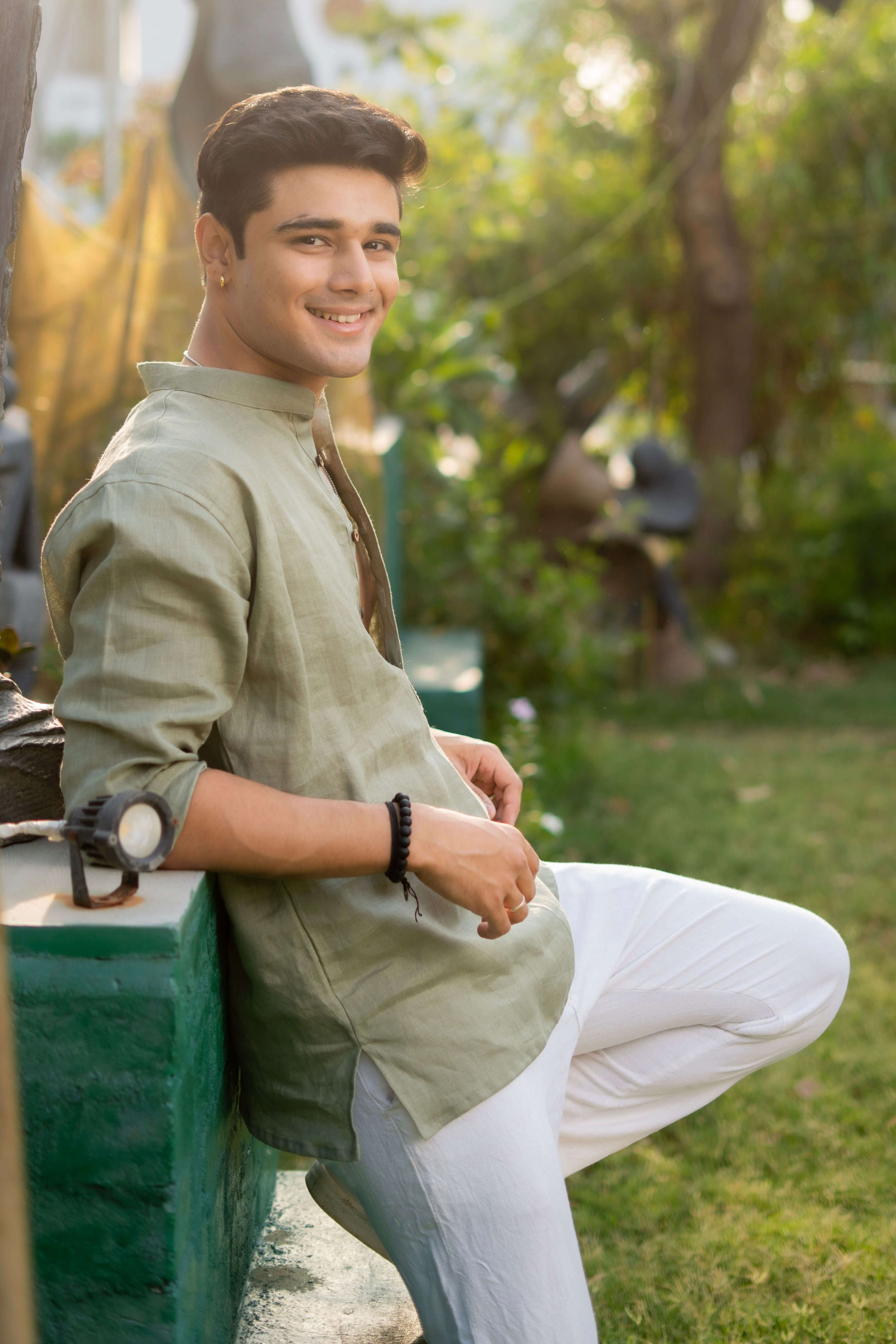 Smiling man wearing a sage green linen short kurta with wooden buttons and white pants, sitting casually on a green wall in a sunlit garden with lush greenery in the background.