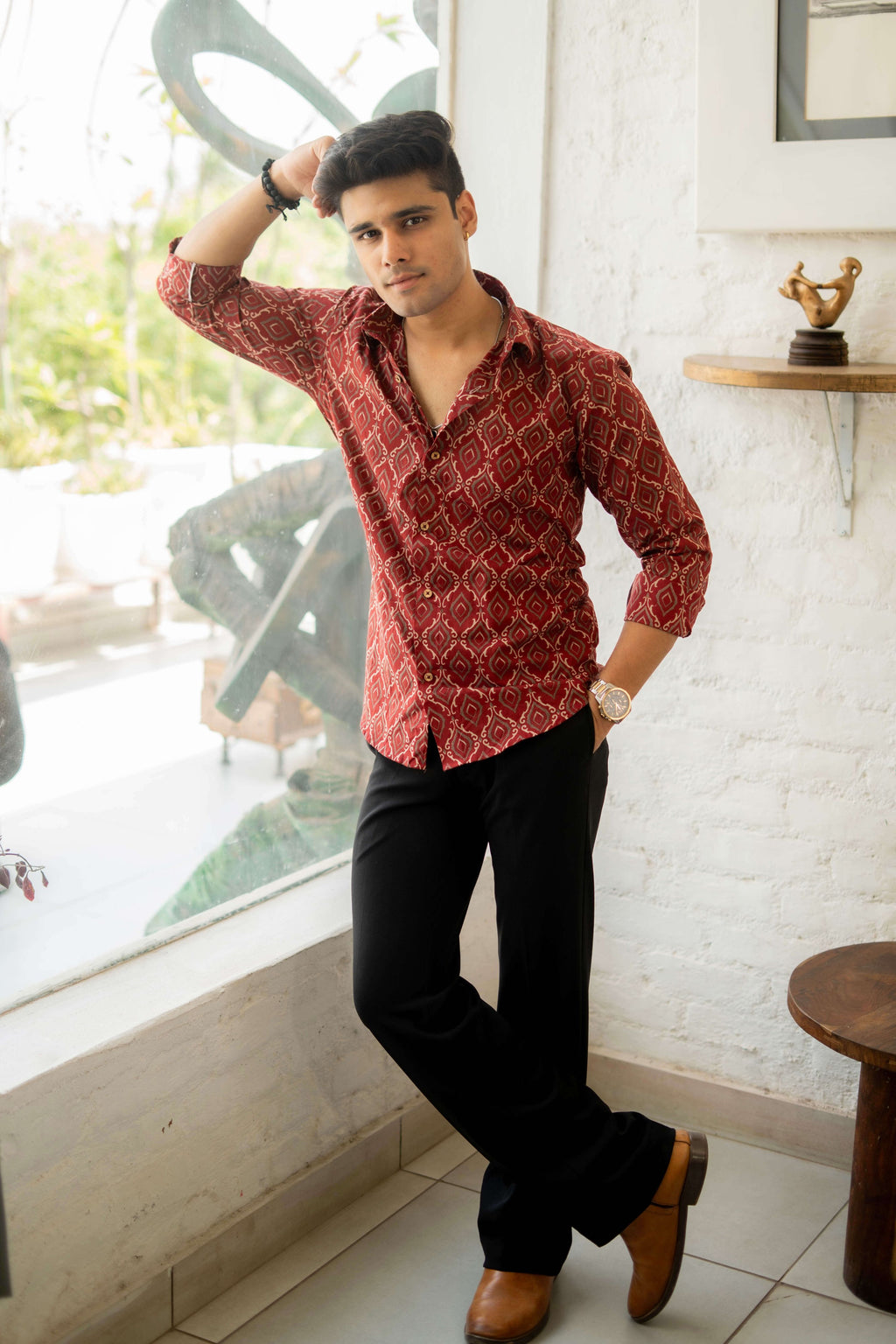 Man wearing a fall red shirt with diamond geometric print, paired with black trousers and tan boots, standing stylishly indoors.