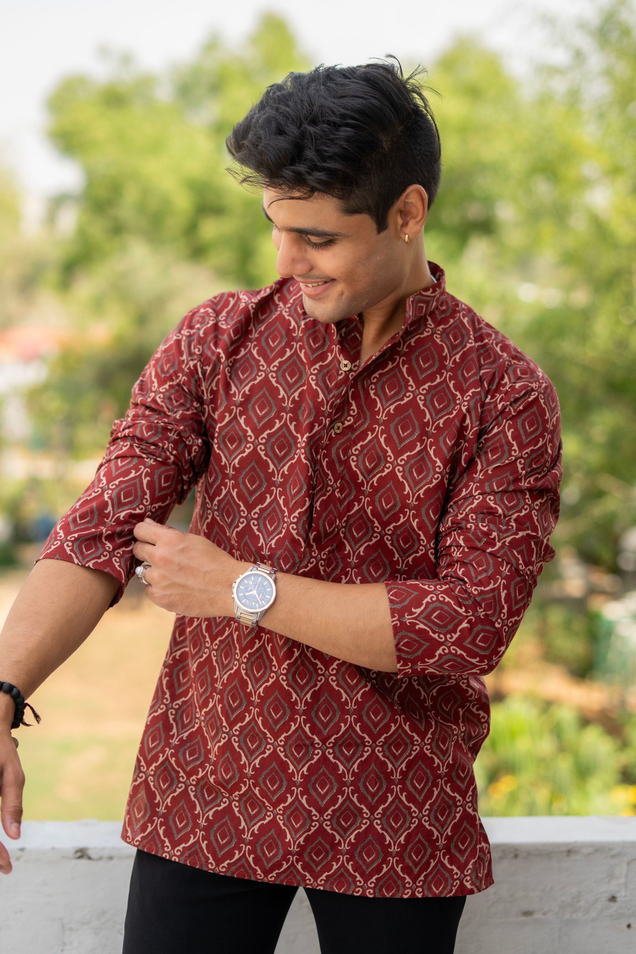 An Indian man in a red geometric print kurta and black pants checks his watch, appearing thoughtful and attentive to time.