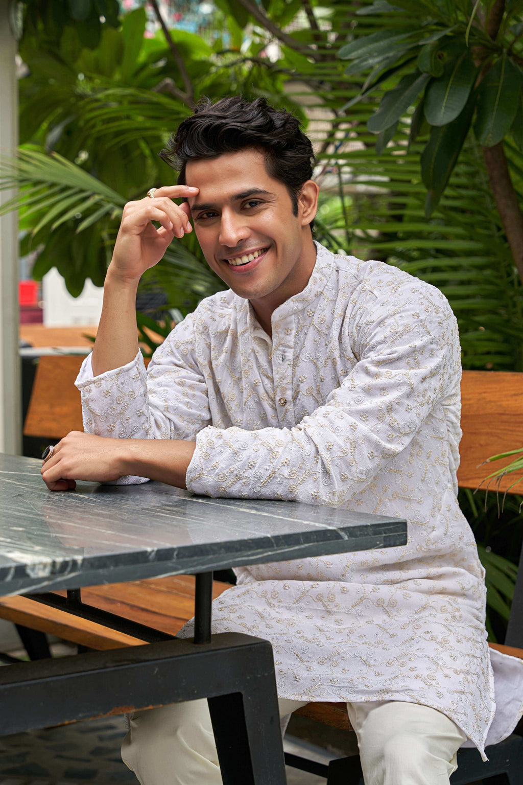 Indian man wearing a white zari work long kurta. sitting on a table.
