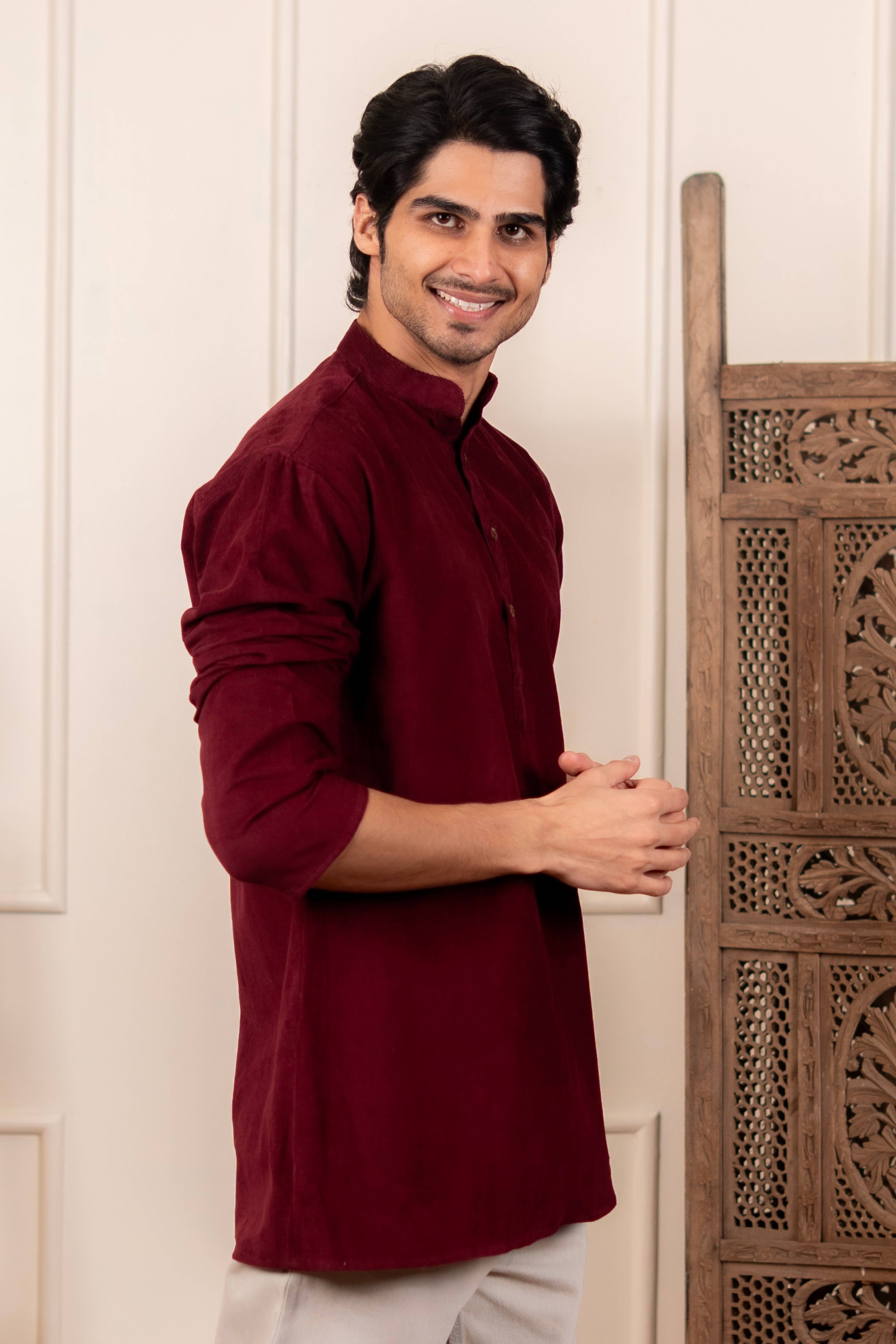 Man wearing a maroon kurta standing next to a wooden decorative piece.