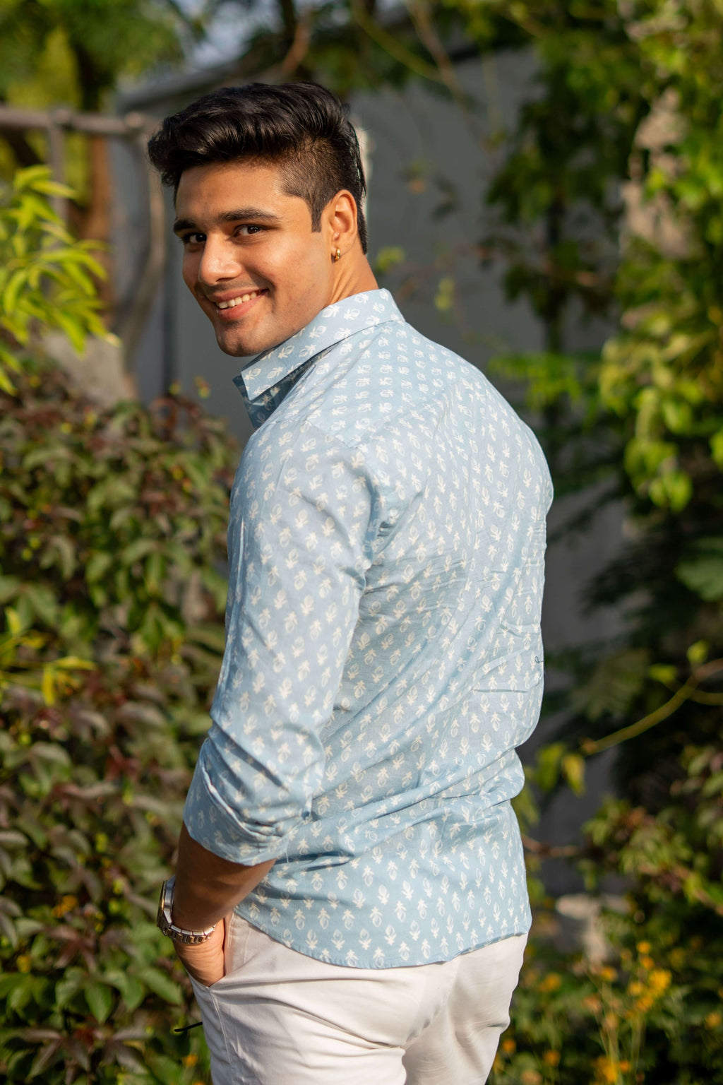  An Indian man wearing a light blue shirt with a white pattern and white pants, posing outdoors with greenery in the background. He accessorizes with a wristwatch and has one hand in his pocket, creating a relaxed and stylish look.