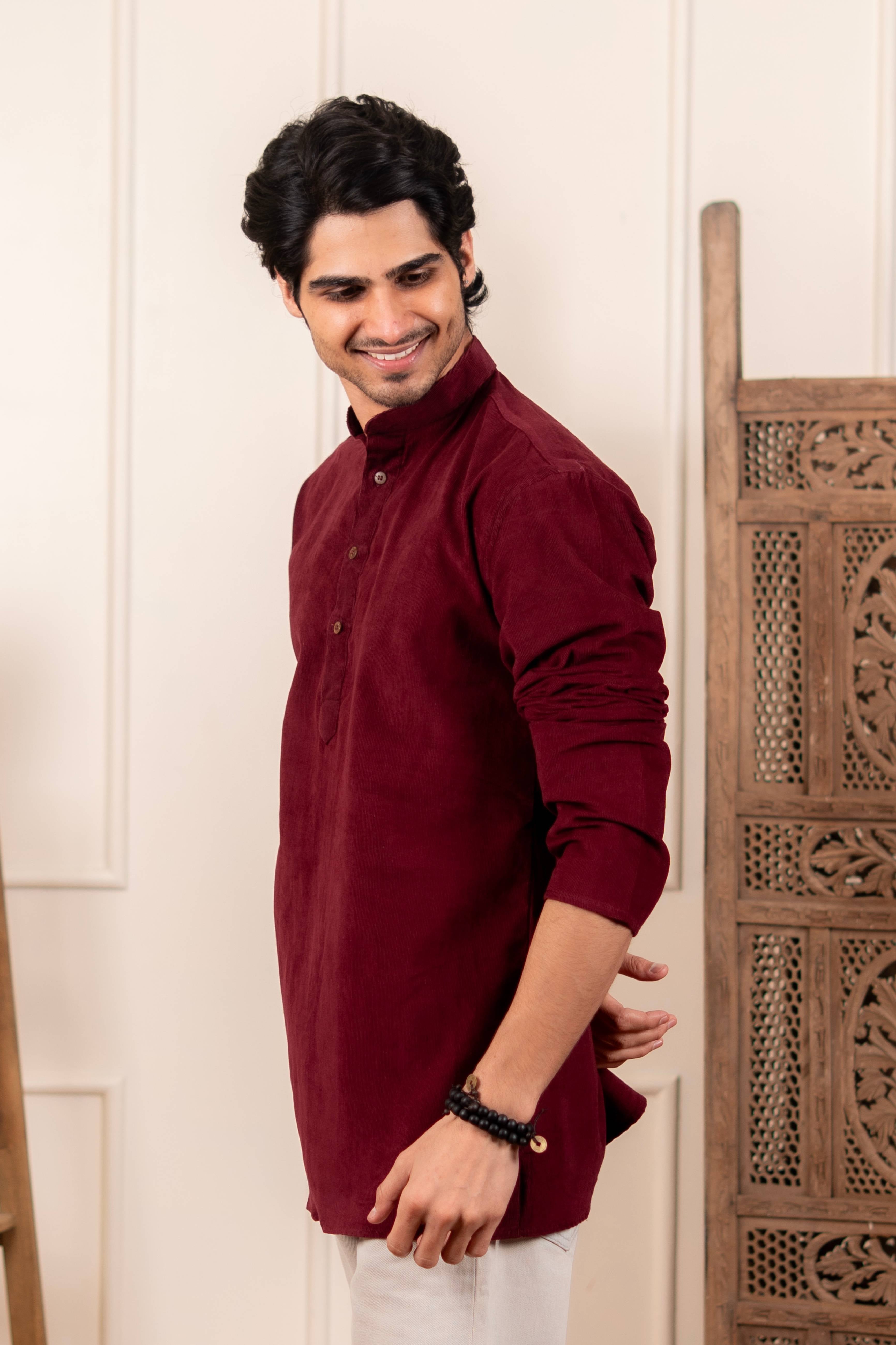 Man wearing a maroon kurta standing next to a wooden chair indoors.