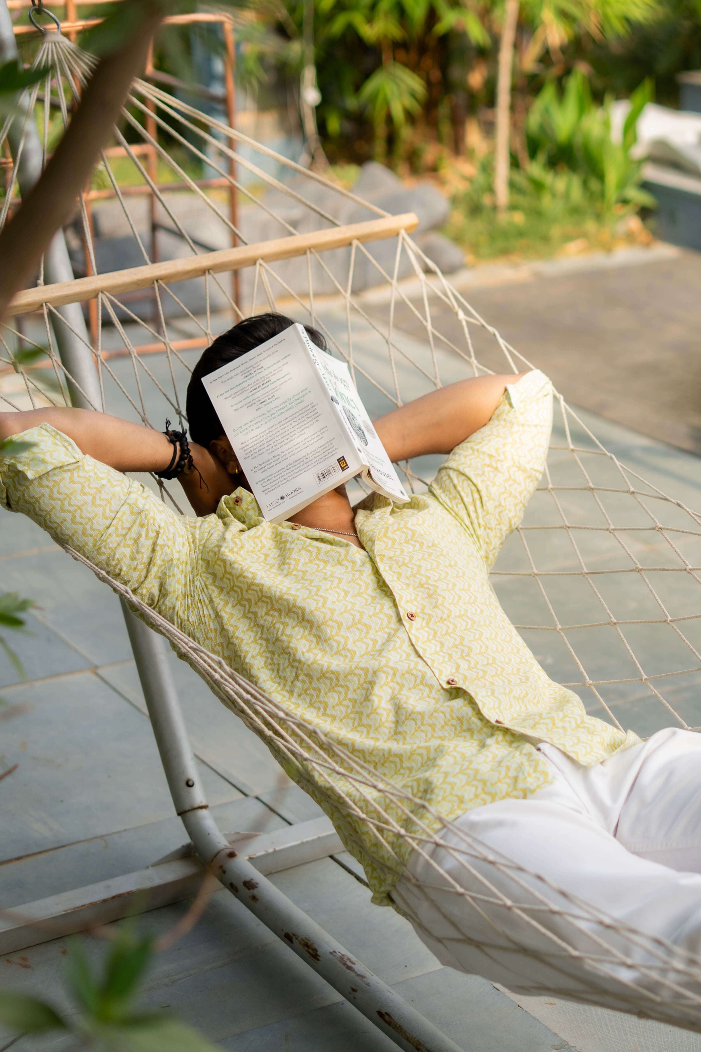 Man relaxing on a hammock wearing a white Kantha work shirt with volt green chevron print, face covered with a book.