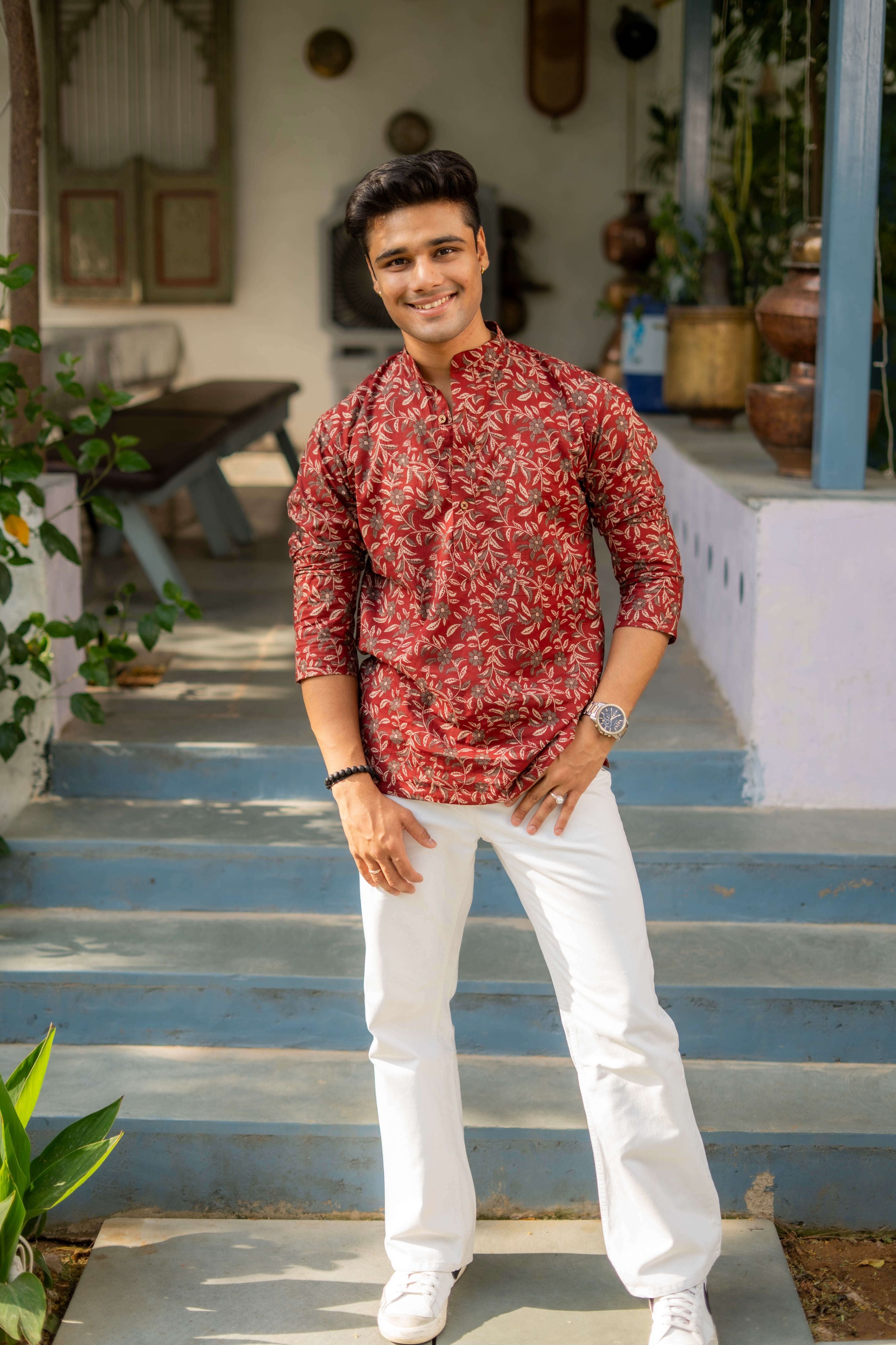 A person wearing a Scarlet Maroon Short Kurta with Floral Print, paired with white pants and white sneakers, standing on blue steps in an outdoor setting with plants and traditional decor in the background.