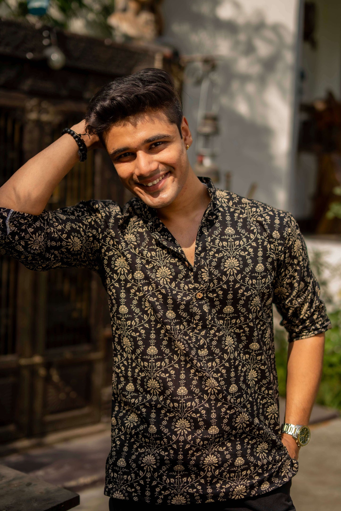 A person wearing a black short kurta with beige Dabu print, paired with white pants, standing in an outdoor setting with traditional decor.