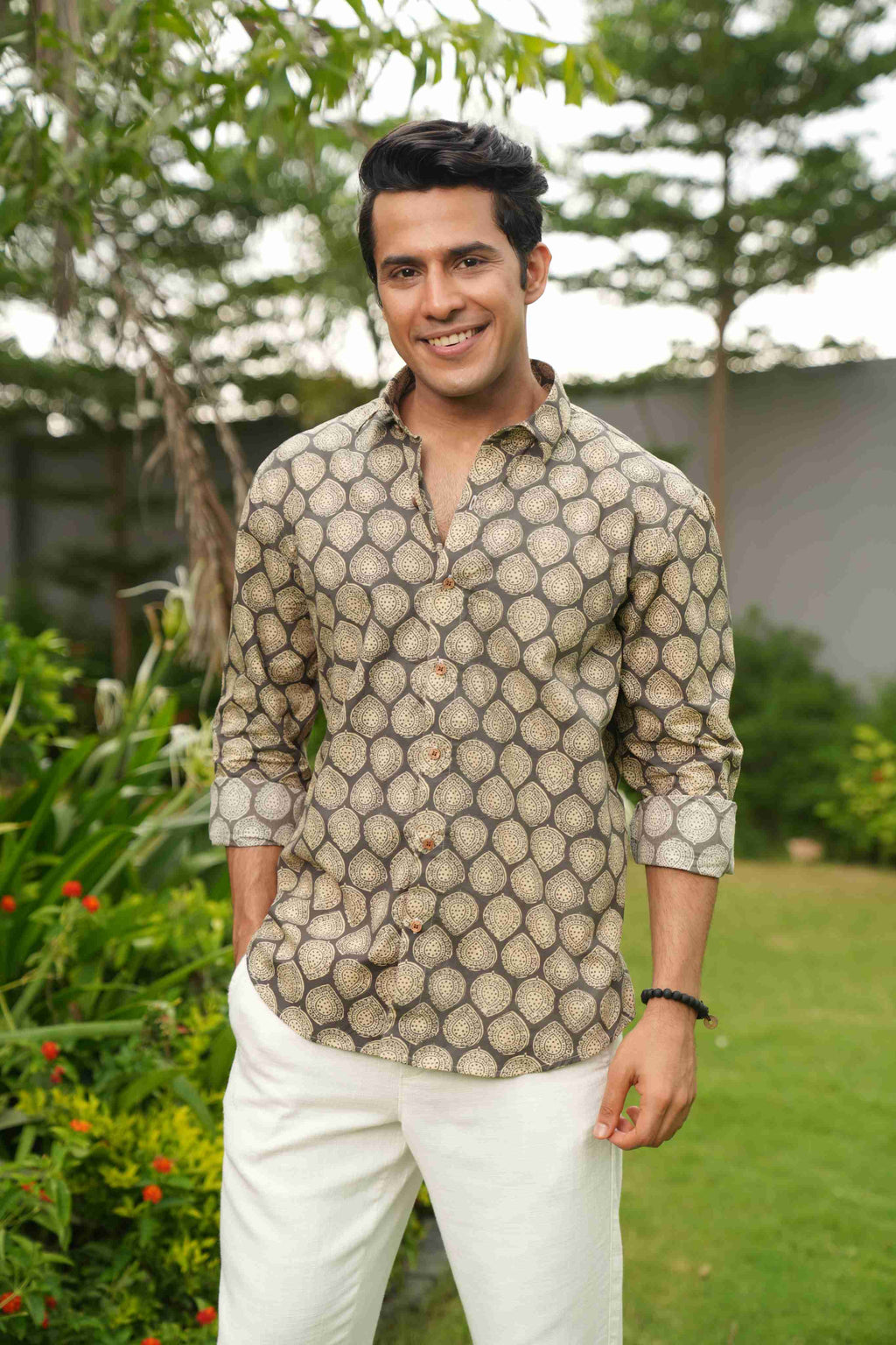 Man wearing a patterned shirt and white pants standing outdoors with greenery in the background