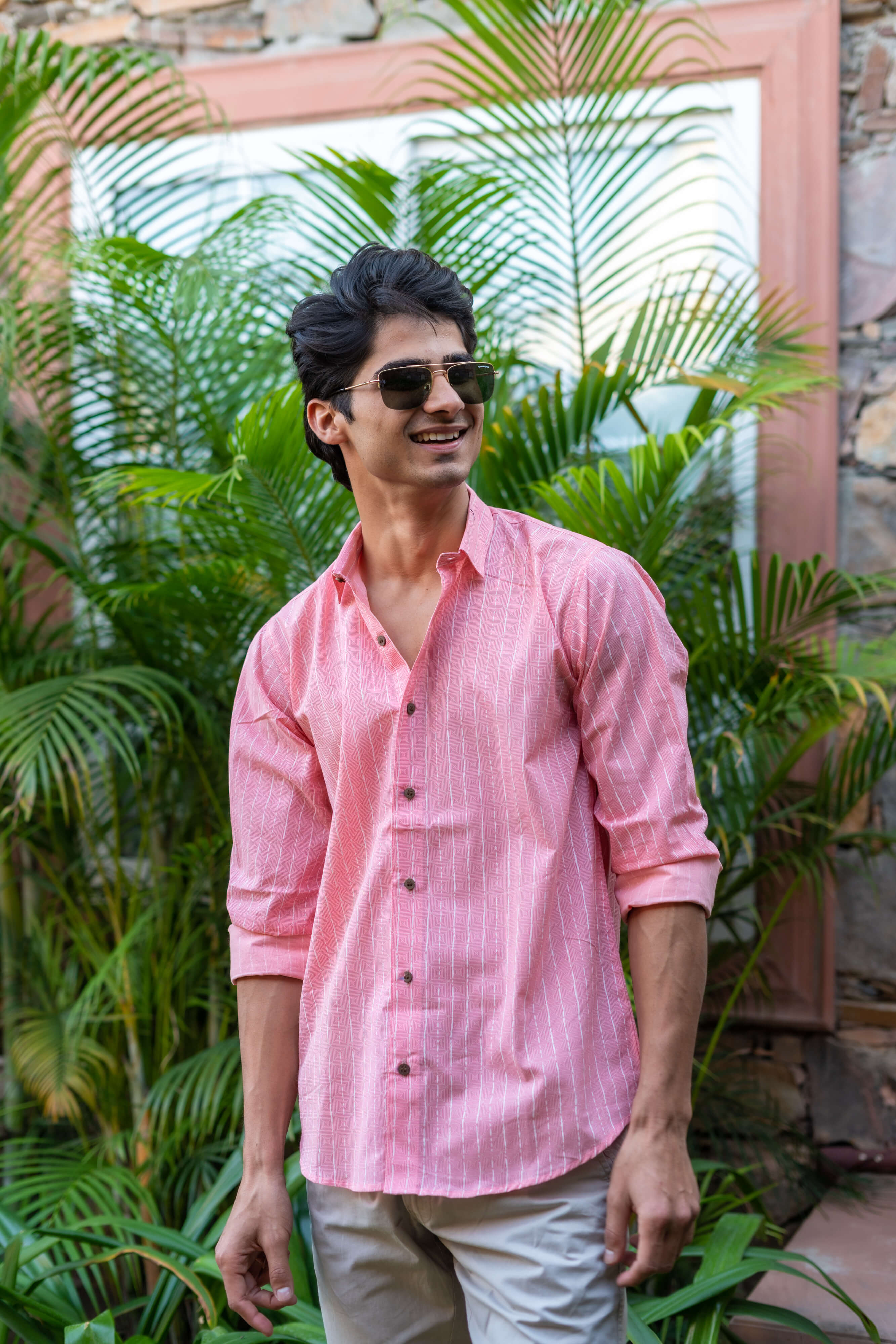 An Indian Man in a Pink Striped Shirt Is Standing in Front of Green Palm Leaves, Wearing Sunglasses and Smiling.