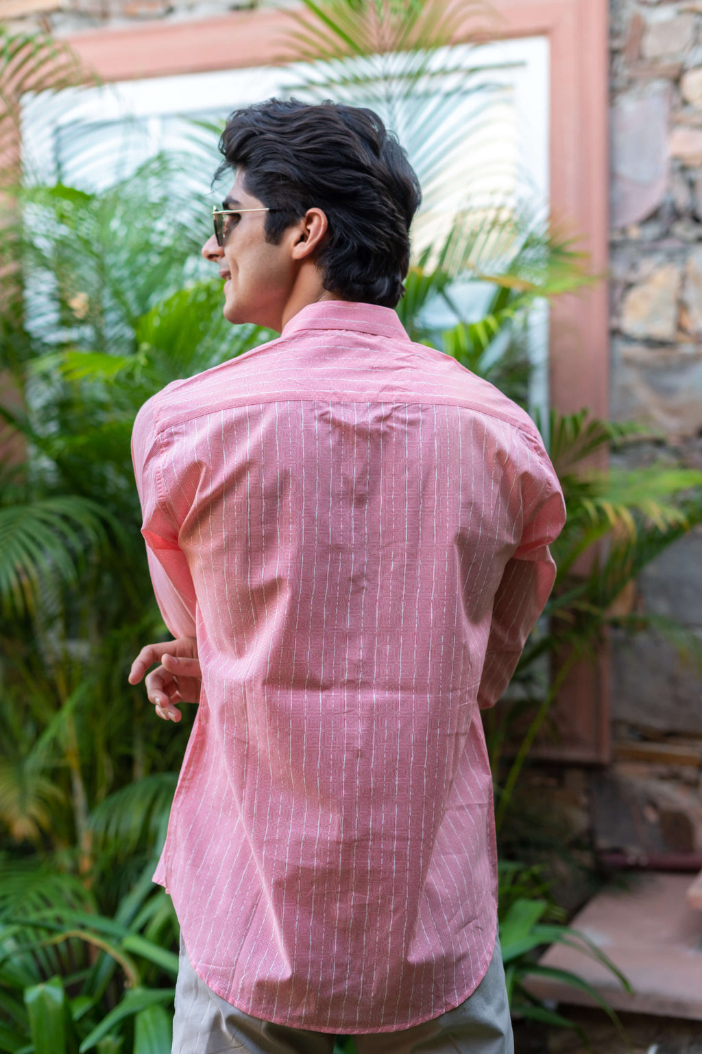 back view of An Indian Man in a Pink Striped Shirt Is Standing in Front of Green Palm Leaves, Wearing Sunglasses and Smiling.