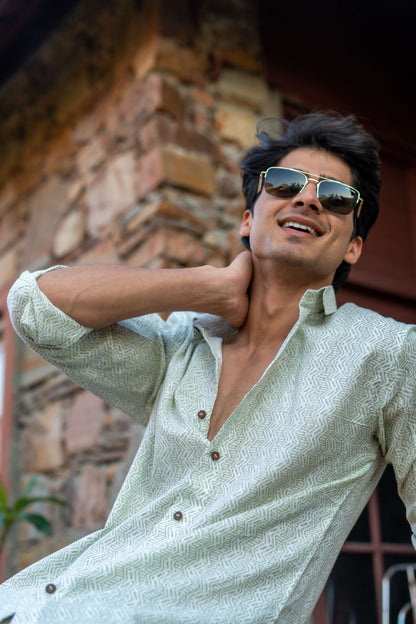 Indian Man Wearing a White Tribal Print Shirt and Black Sun Glasses, Sitting in Front of a Wall and a Plant.