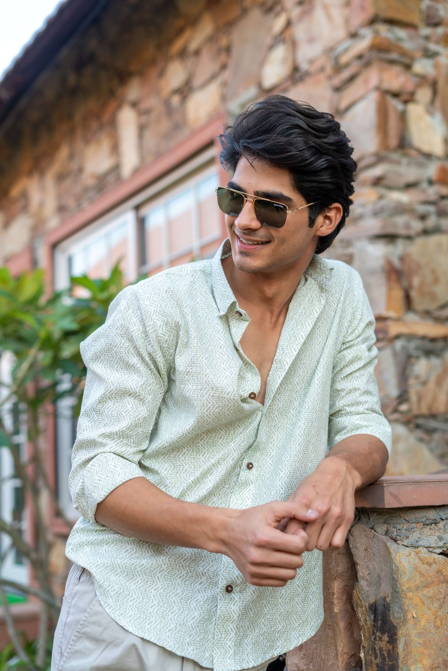 Indian man wearing a white tribal print shirt, standing in front of a wall and a plant.