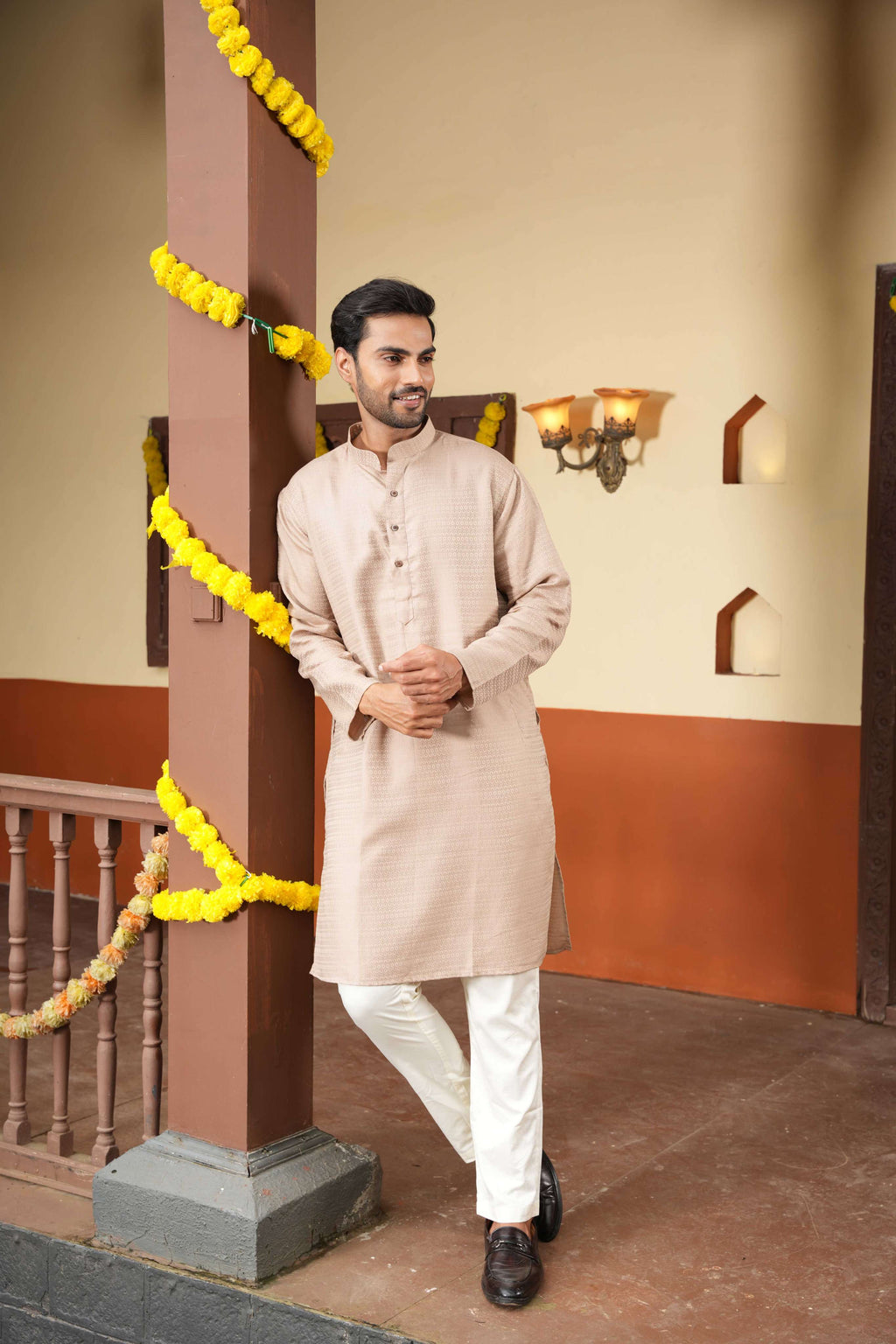 Man in a beige kurta with white pants standing next to a decorated pillar indoors.