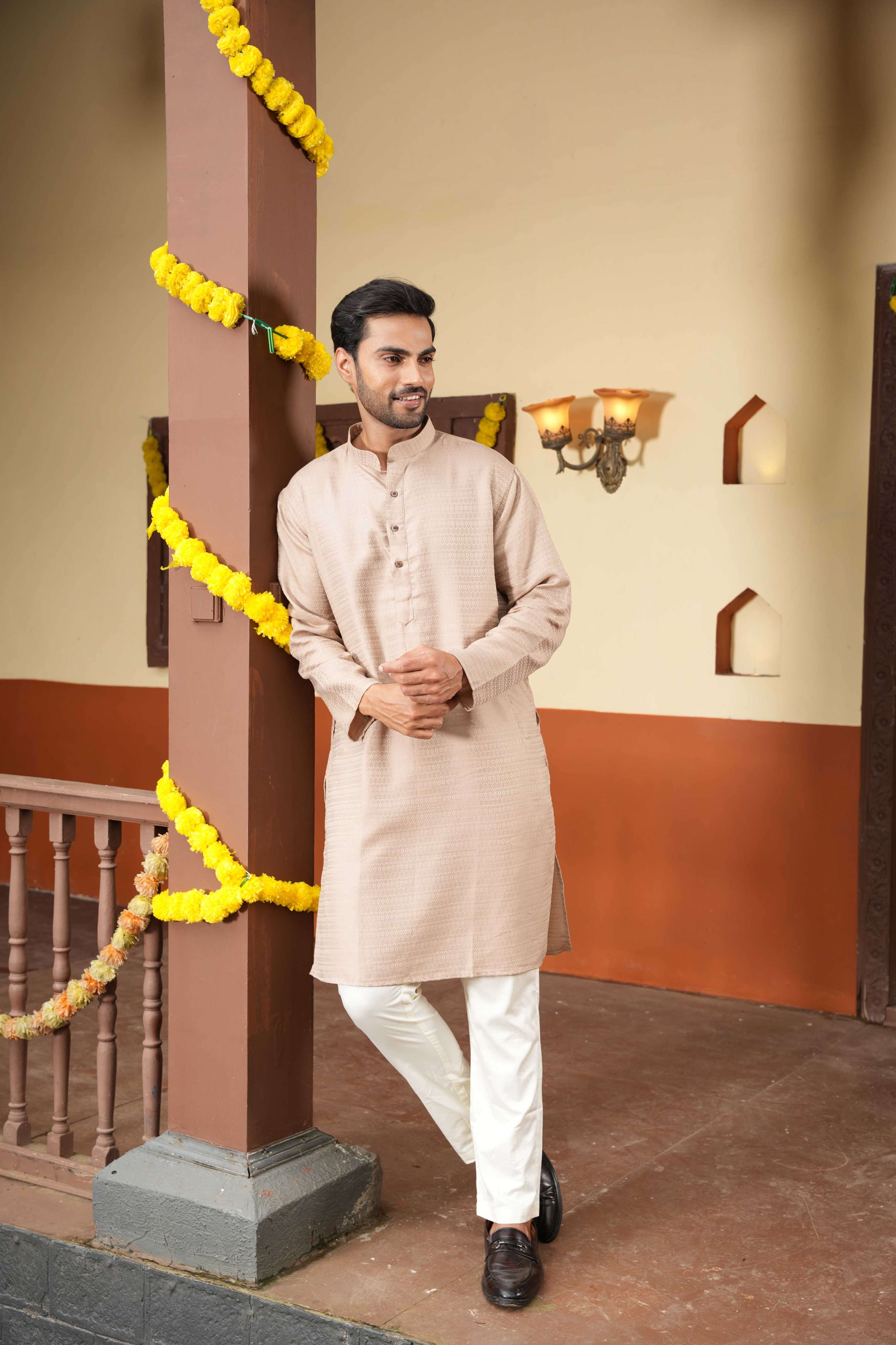 Man in a beige kurta with white pants standing next to a decorated pillar indoors.