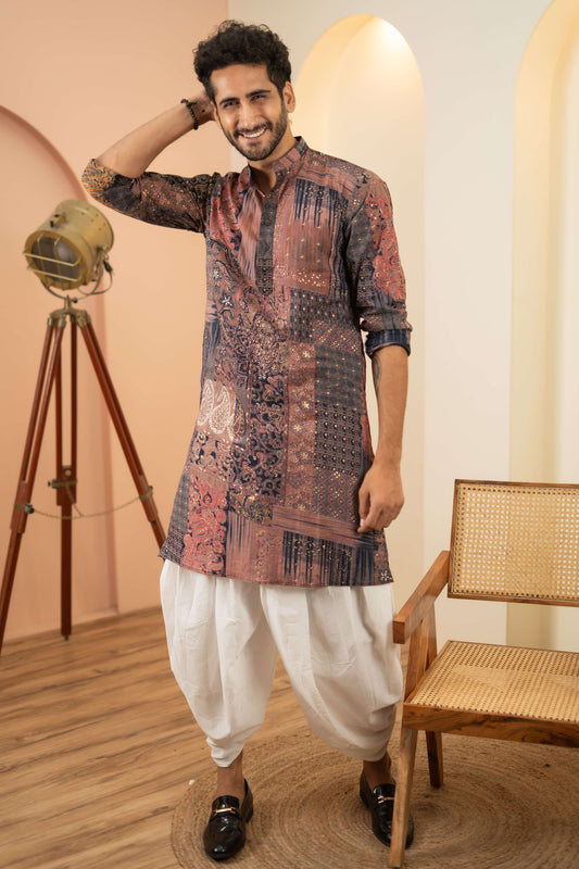 Indian man in a patchwork kurta, white trousers, and black shoes, standing next to a chair.