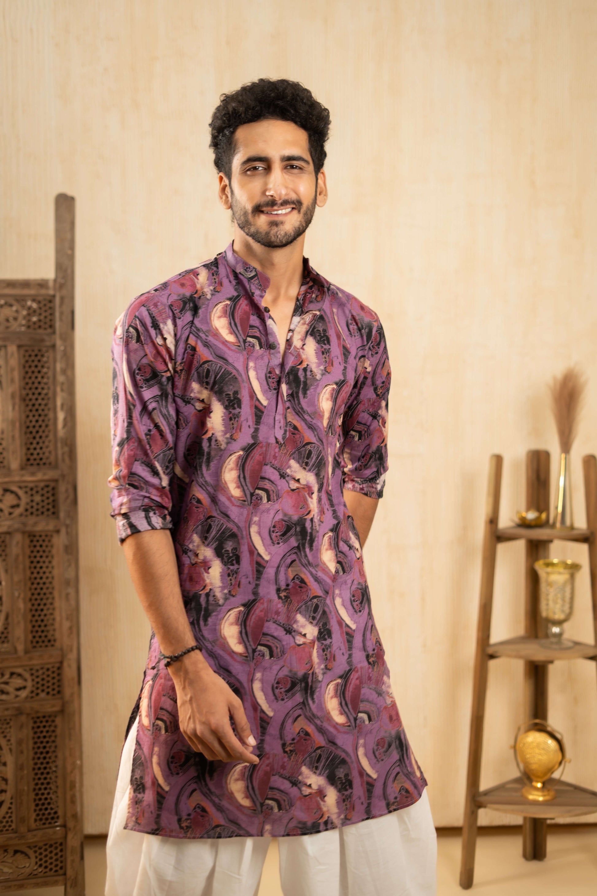 Indian man in a purple kurta and dhoti, standing inside a decorated room.