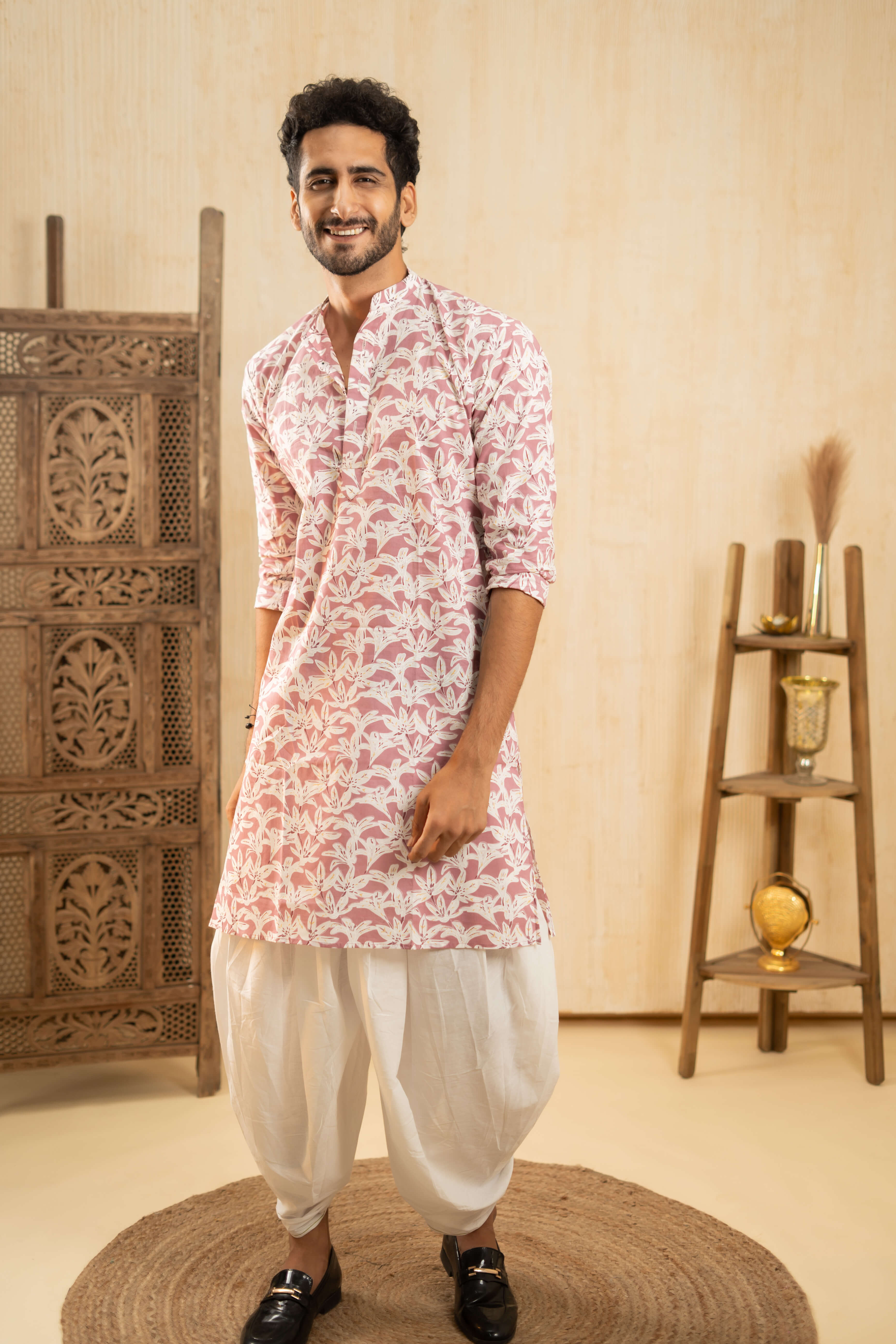 A person wearing a pink and white floral patchwork kurta and white dhoti pants stands on a round mat. The background includes a wooden carved screen and a wooden shelf with decorative items.