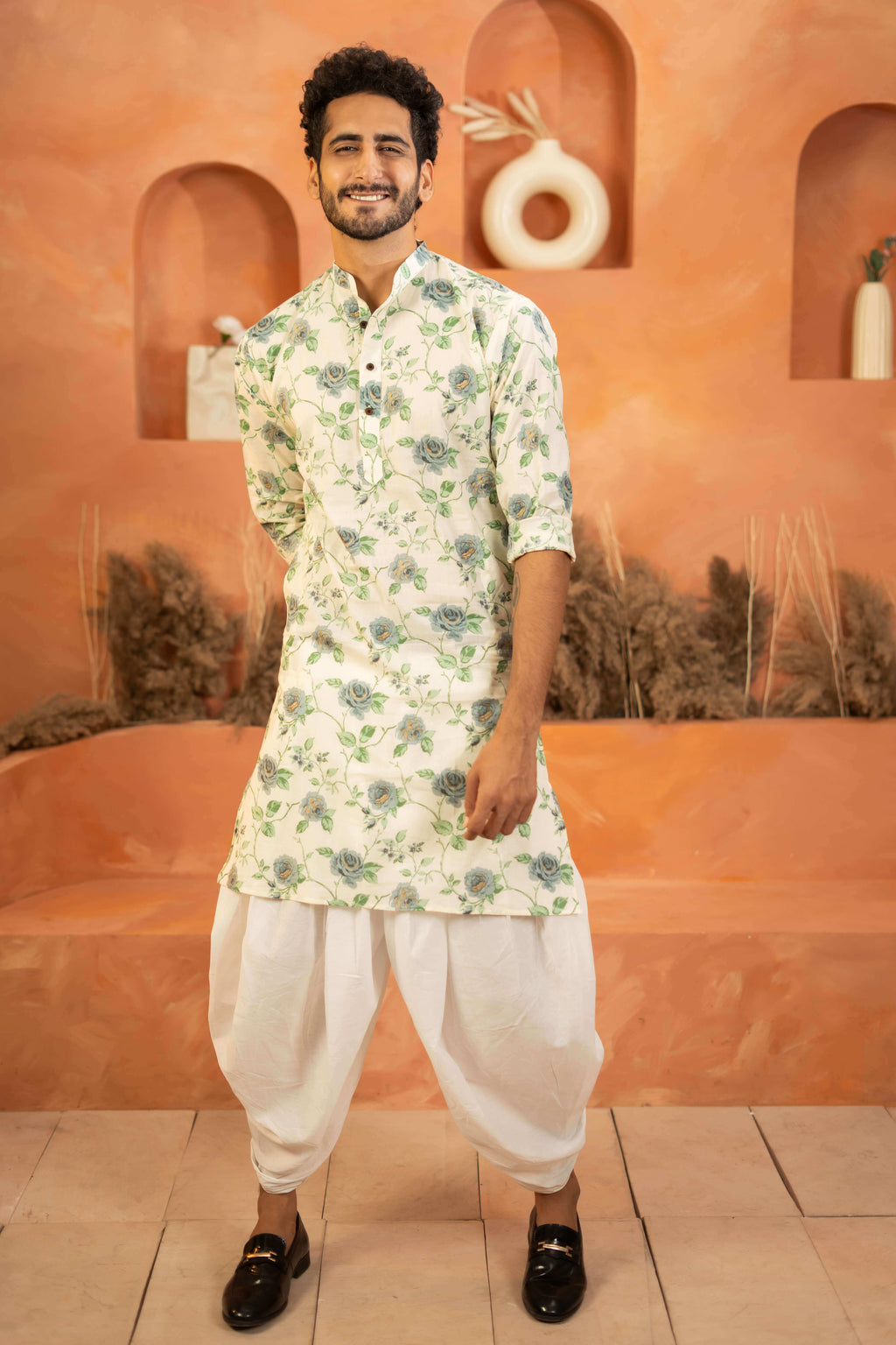 Indian men wearing a long kurta with a floral print. He is pairing this kurta with a white colored dhoti and black formal shoes. 