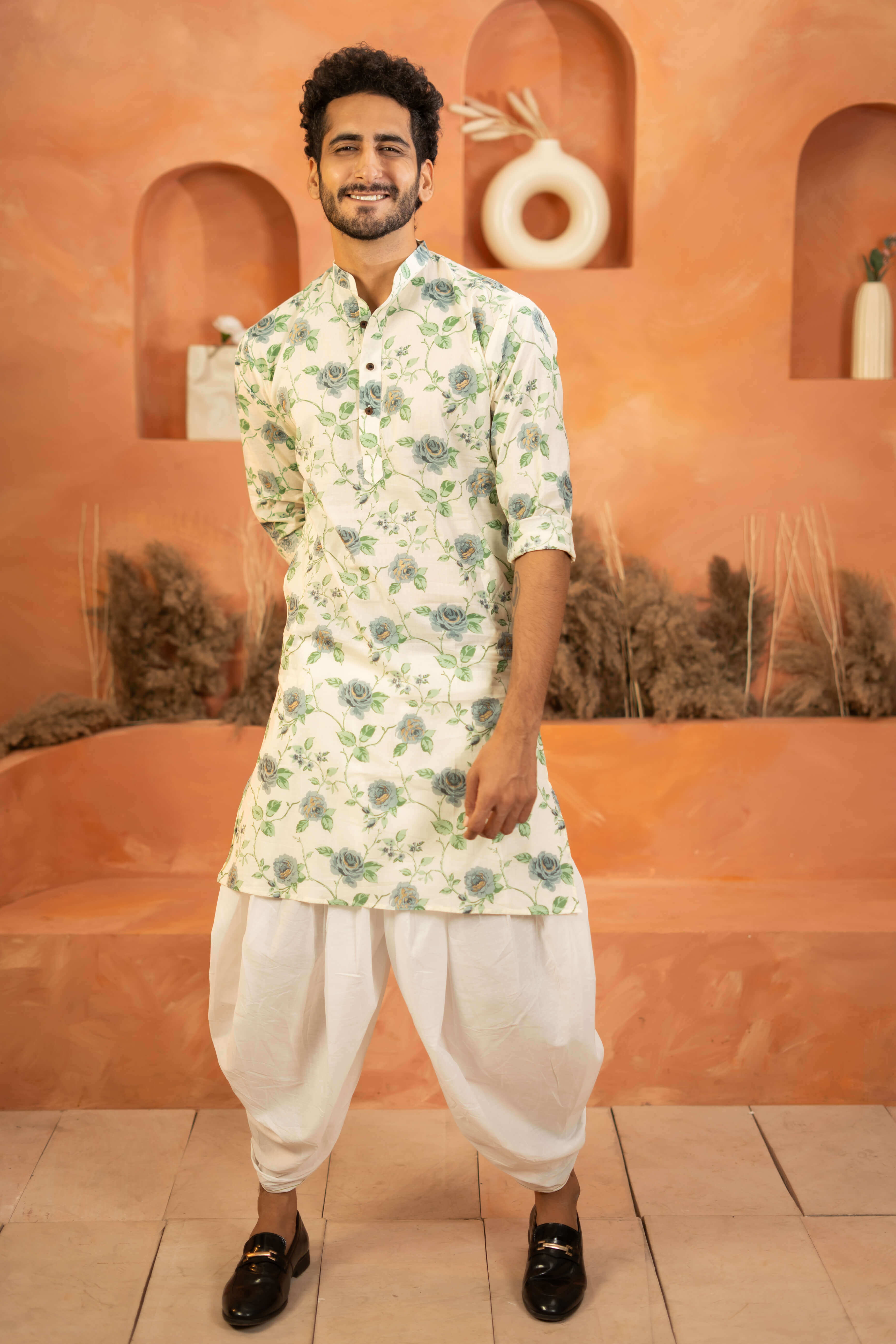 Indian men wearing a long kurta with a floral print. He is pairing this kurta with a white colored dhoti and black formal shoes. 
