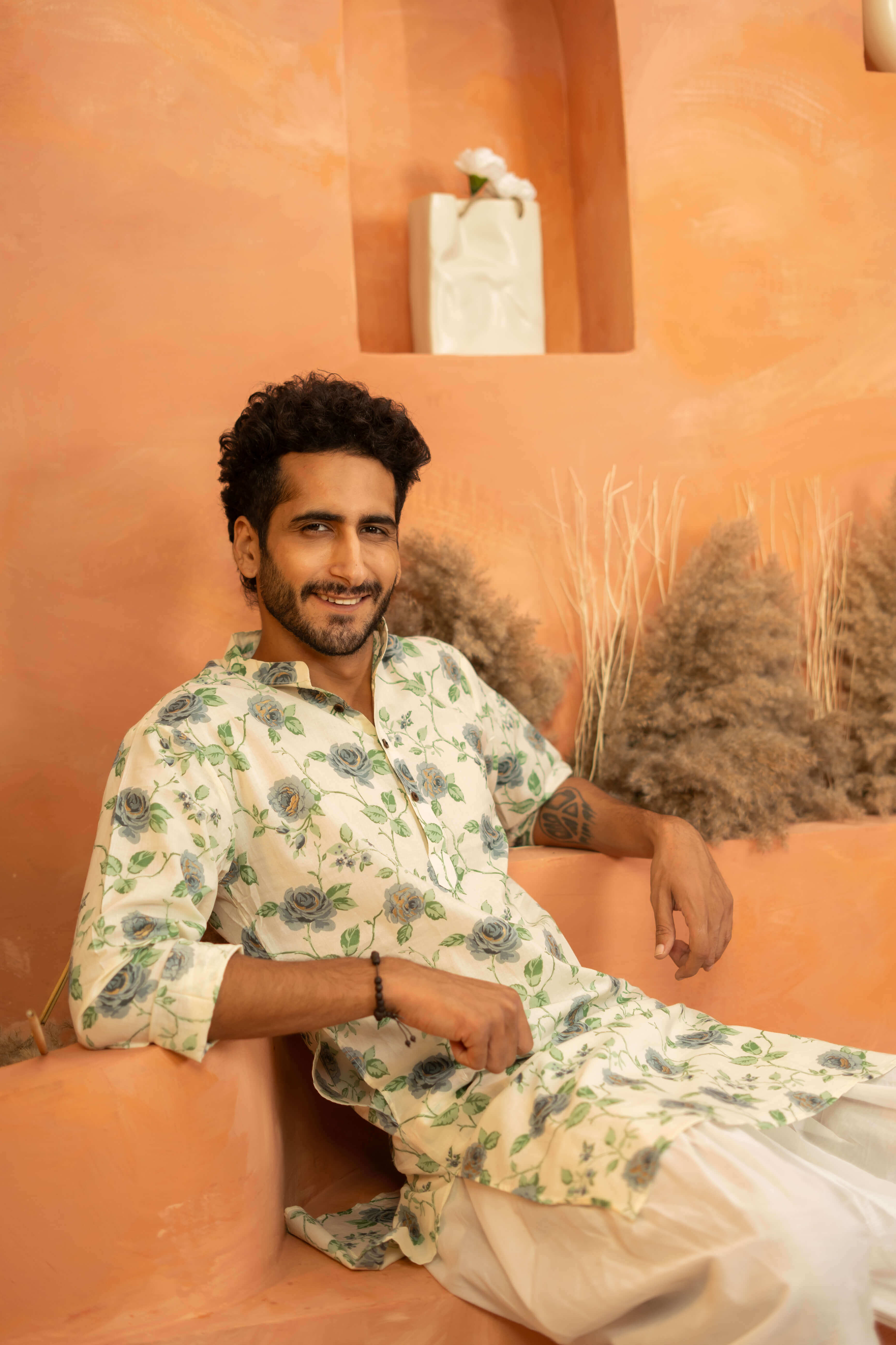 An indian man sitting on an orange surface wearing a white floral kurta and white pants.