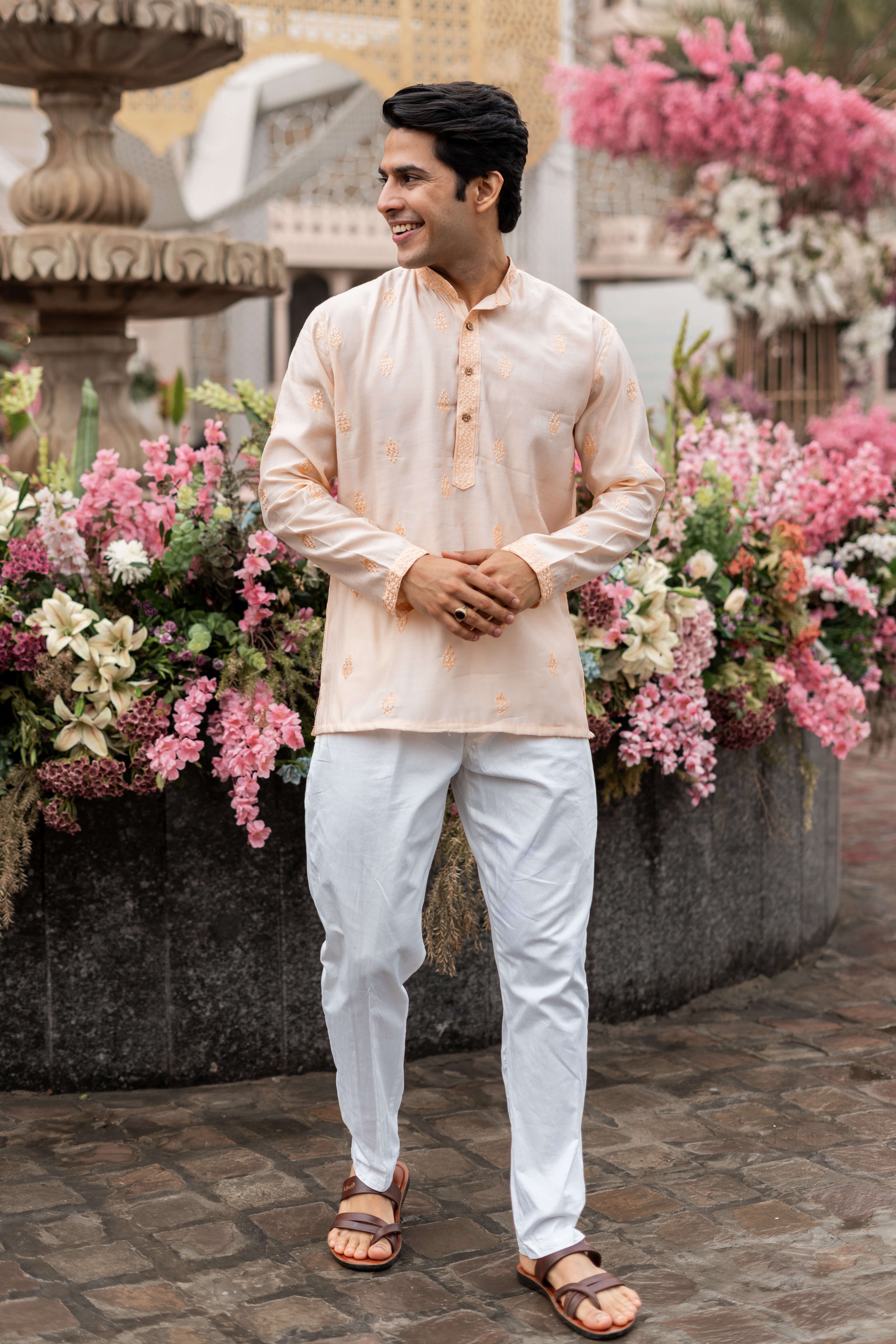 A man wearing a pink short kurta with butti embroidery and white pants stands in front of a decorative fountain.