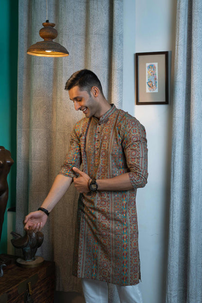 Man wearing an intricately patterned brown kurta with white trousers, smiling while interacting with a decorative sculpture in an elegant indoor setting.