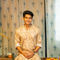 an Indian man smiling and wearing a brown color long kurta with an off-white floral print sitting on a table 